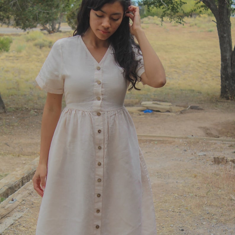May include: A light beige linen dress with a button-up front and short sleeves. The dress has a gathered waist and a flowy skirt.