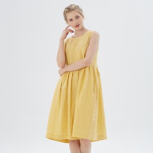 May include: A sleeveless, knee-length yellow dress. The dress features a round neckline, a gathered waist, and pockets. The fabric appears to be linen or a similar lightweight material. The model is wearing the dress in a studio setting with a white background.