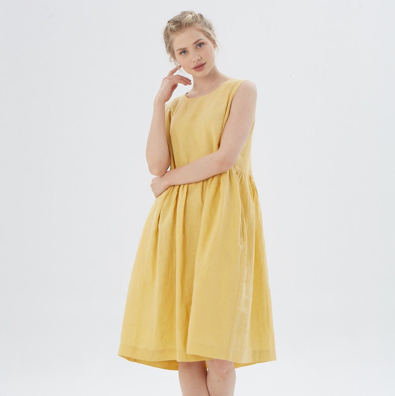 May include: A sleeveless, knee-length yellow dress. The dress features a round neckline, a gathered waist, and pockets. The fabric appears to be linen or a similar lightweight material. The model is wearing the dress in a studio setting with a white background.