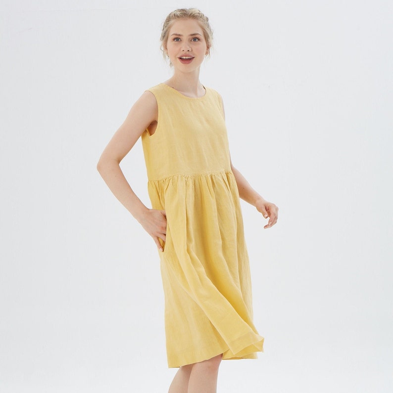 May include: A sleeveless, knee-length yellow dress with a gathered waist and side pockets. The dress is made of a lightweight fabric and has a simple, casual design. The model is wearing the dress in a studio setting.