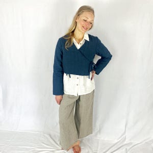 May include: A teal blue wrap jacket with long sleeves and a tie at the waist. The jacket is worn over a white button-down shirt and beige wide-leg pants.