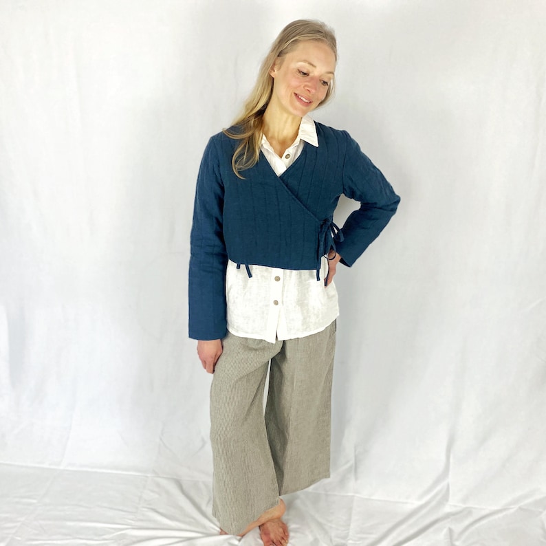 May include: A teal blue wrap jacket with long sleeves and a tie at the waist. The jacket is worn over a white button-down shirt and beige wide-leg pants.