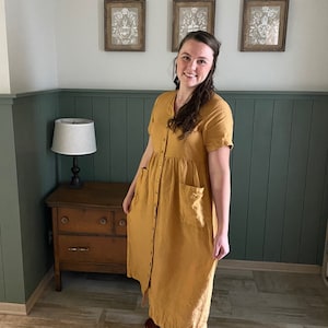 May include: A woman wearing a long, mustard yellow linen dress with a button-up front and pockets. She is standing in a room with green wainscoting and a wooden dresser.
