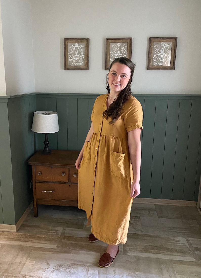 May include: A woman wearing a long, mustard yellow linen dress with a button-up front and pockets. She is standing in a room with green wainscoting and a wooden dresser.