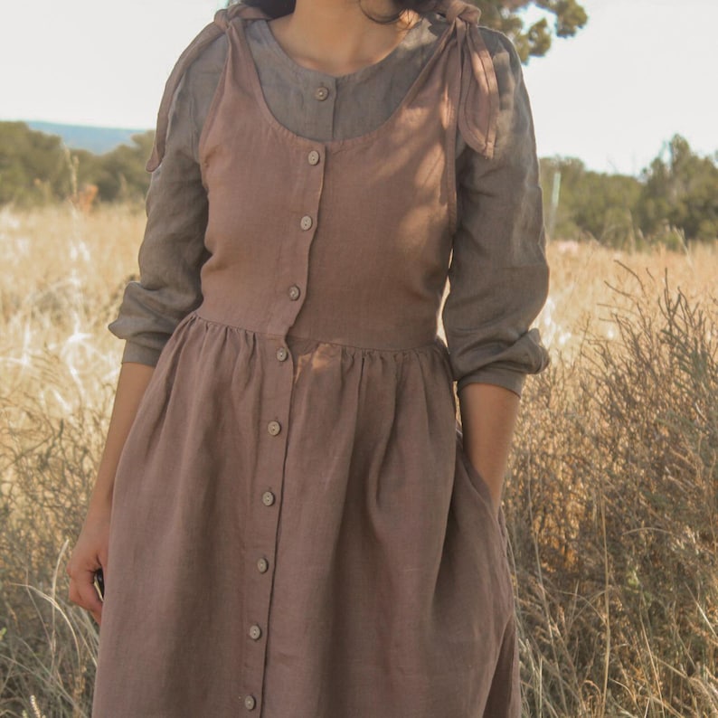 May include: A button-down, dusty rose-colored dress with a scoop neckline and a gathered skirt. The dress is layered over a long-sleeved, gray-green blouse. The dress has a vintage, prairie-style aesthetic.
