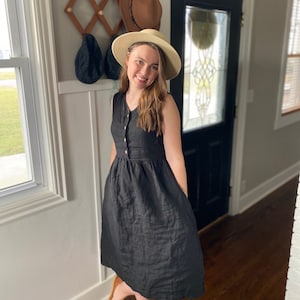 Pode incluir: Uma mulher vestindo um vestido de linho preto com bot&otilde;es na frente e bolsos. Ela est&aacute; usando um chap&eacute;u de palha e tem cabelo castanho longo.