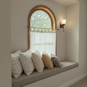 a cozy and inviting window seat with a white curtain and a wooden frame, adorned with four pillows in various colors and patterns.