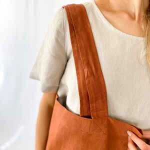May include: A rust-colored linen tote bag with double straps. The bag is made of a natural fabric and has a simple, minimalist design.
