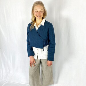 May include: A blue quilted wrap jacket with a tie closure. The jacket is worn over a white button-down shirt and striped pants.