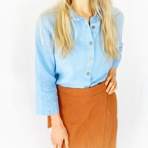 May include: A light blue linen shirt with a button-up front and long sleeves. The shirt is paired with a rust-colored linen wrap skirt.