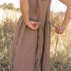 May include: A brown linen dress with a tie waist. The dress has a simple design with a gathered waistline and a flowing skirt. The dress is being tied by the wearer. The background is a field of dry grass.