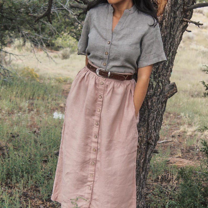 May include: A woman wearing a gray striped button-up shirt with a brown leather belt and a long, dusty pink button-up skirt.