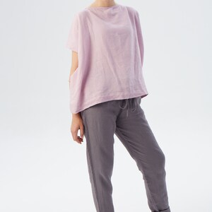 May include: A woman is wearing a loose-fitting, pale pink linen top with short sleeves and a pair of dark gray linen pants. The pants have a drawstring waist and are cuffed at the ankles.