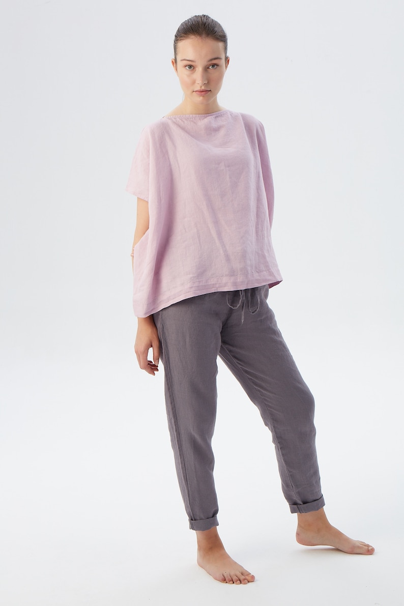 May include: A woman is wearing a loose-fitting, pale pink linen top with short sleeves and a pair of dark gray linen pants. The pants have a drawstring waist and are cuffed at the ankles.