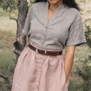 May include: A woman wearing a gray striped linen shirt with a brown leather belt and a pink button-down skirt.