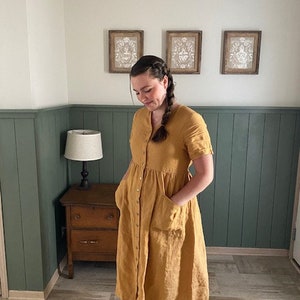 May include: A woman wearing a mustard yellow linen dress with a button-up front and pockets. The dress has a gathered skirt and short sleeves.