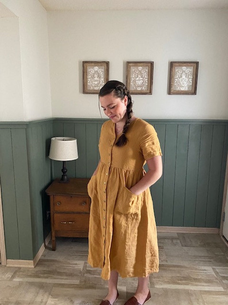 May include: A woman wearing a mustard yellow linen dress with a button-up front and pockets. The dress has a gathered skirt and short sleeves.