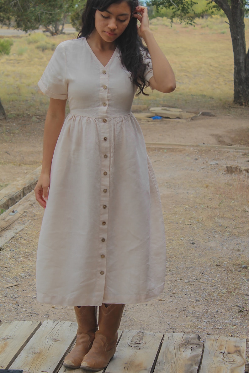 May include: A woman wearing a long, light pink linen dress with a button-down front and short sleeves. She is wearing brown cowboy boots and standing on a wooden platform.