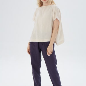 May include: A woman is wearing a loose-fitting, off-white linen top with short sleeves and a pair of dark blue linen pants.