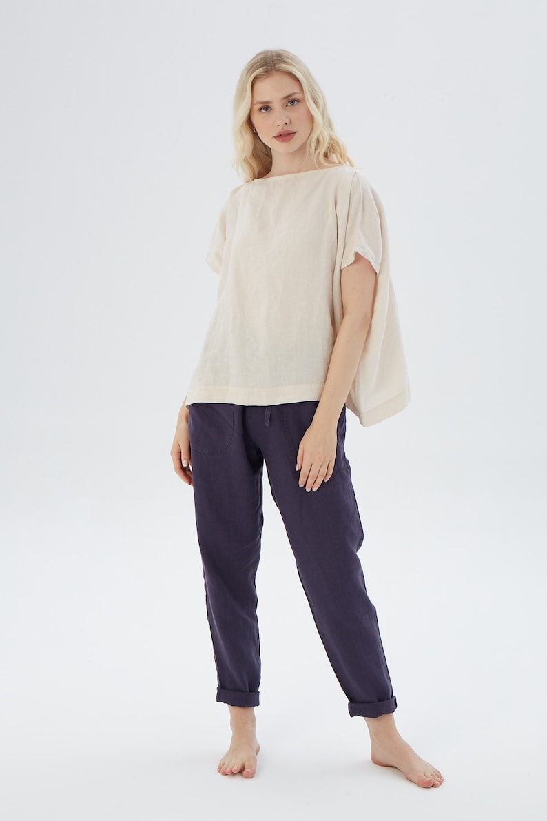 May include: A woman is wearing a loose-fitting, off-white linen top with short sleeves and a pair of dark blue linen pants.