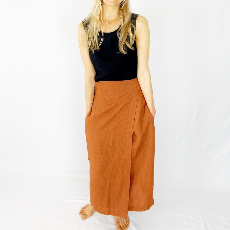 May include: A rust-colored linen wrap skirt with a long, flowing silhouette. The skirt has a simple design with a single wrap and a deep front pocket on each side.