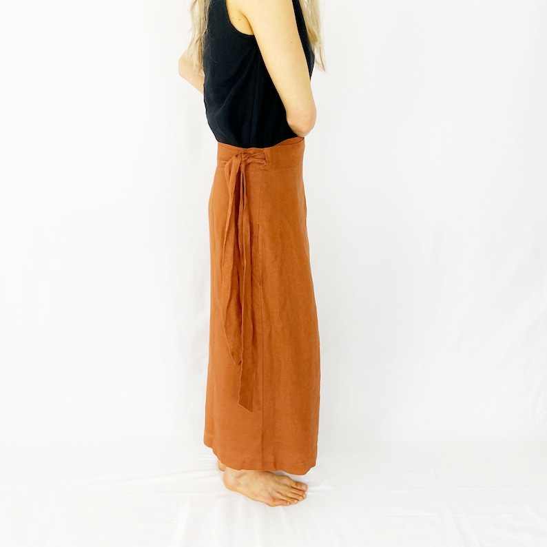 May include: A rust-colored linen wrap skirt with a long, flowing silhouette. The skirt is tied at the waist with a matching fabric belt.