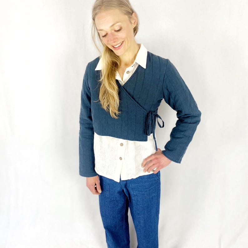 May include: A dark blue quilted wrap jacket with long sleeves and a tie closure at the waist. The jacket is worn over a white button-down shirt and blue jeans.