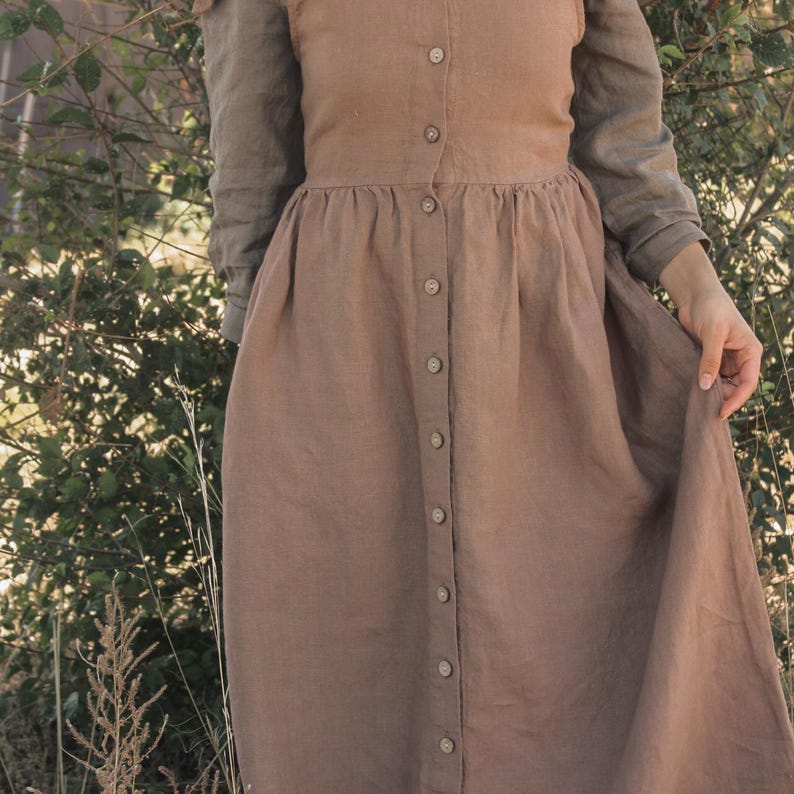 May include: A brown linen dress with long sleeves and a button-down front. The dress has a fitted bodice and a flowing skirt. The sleeves are a lighter shade of brown. The dress is made of a soft, natural fabric.