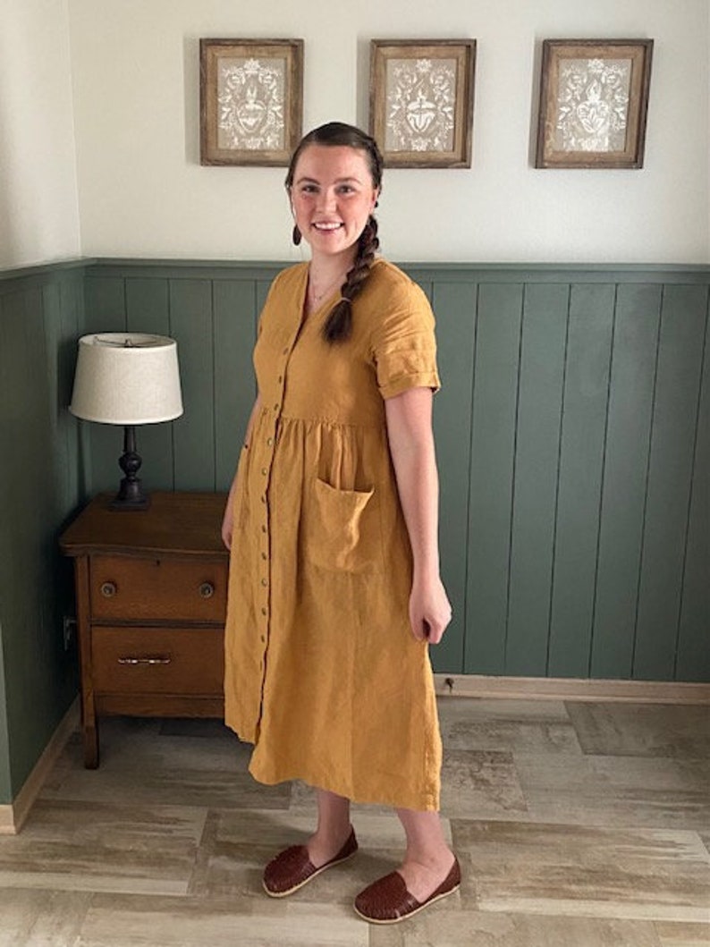 May include: A woman wearing a mustard yellow linen dress with a button-down front and a patch pocket. She is wearing brown leather sandals.