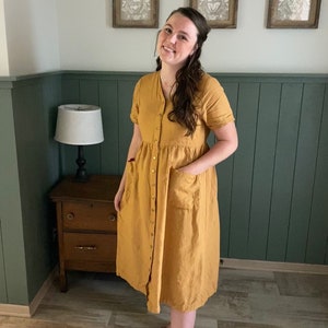 May include: A woman wearing a mustard yellow button-down dress with short sleeves and pockets. She is standing in front of a green wainscot wall with three framed pictures above.