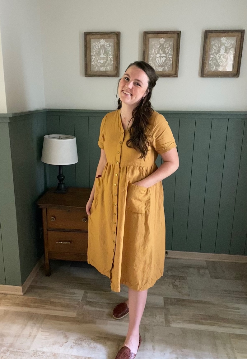 May include: A woman wearing a mustard yellow button-down dress with short sleeves and pockets. She is standing in front of a green wainscot wall with three framed pictures above.