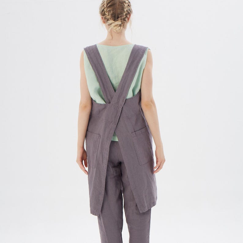 May include: A long, gray linen apron with a criss-cross back strap. The apron has two large pockets on the front.