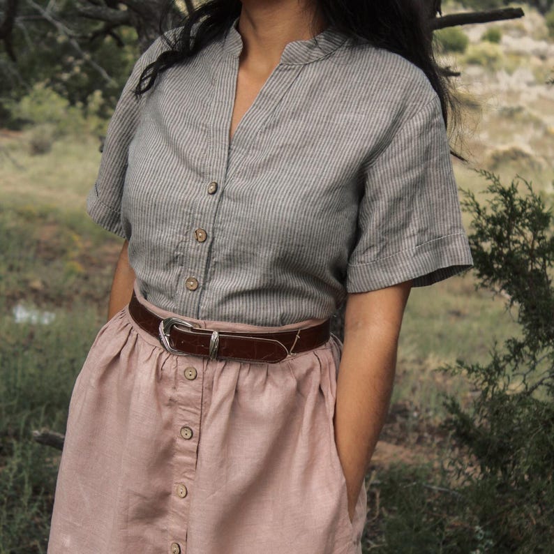 May include: A gray and white striped linen shirt with a button-up front and short sleeves. The shirt is paired with a blush pink skirt with a button-up front and a brown leather belt.