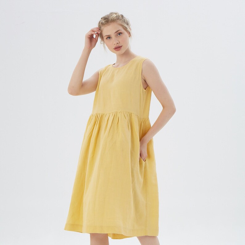 May include: A sleeveless, yellow linen dress with a gathered waist and pockets. The dress falls to the mid-calf and has a simple, round neckline. The model is wearing the dress in a studio setting with a white background.