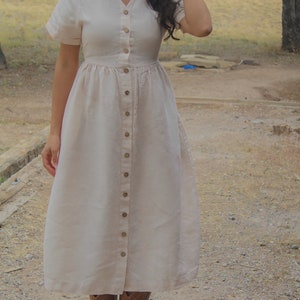 May include: A woman wearing a long, light pink linen dress with a button-down front and short sleeves. She is wearing brown cowboy boots and standing on a wooden platform.