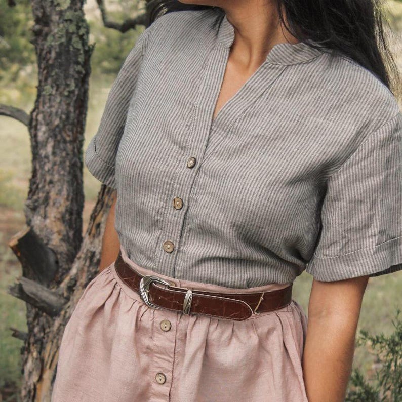 May include: A striped, light gray and white button-up shirt with a v-neck collar. The shirt is tucked into a blush pink skirt with a brown leather belt.