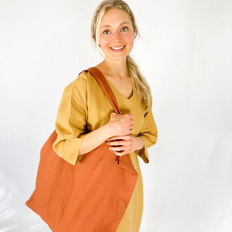 May include: A woman wearing a mustard yellow linen dress is holding a large, rust-colored tote bag.