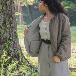 May include: A woman wearing a light olive green quilted jacket with a round collar and a button-down shirt. The jacket has a striped interior and a pocket. She is also wearing a striped skirt and a brown belt. The scene is set outdoors in a forest.
