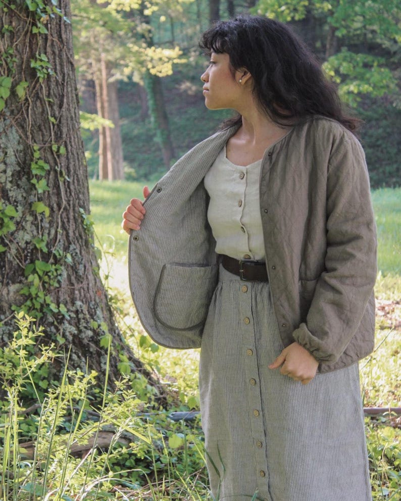 May include: A woman wearing a light olive green quilted jacket with a round collar and a button-down shirt. The jacket has a striped interior and a pocket. She is also wearing a striped skirt and a brown belt. The scene is set outdoors in a forest.