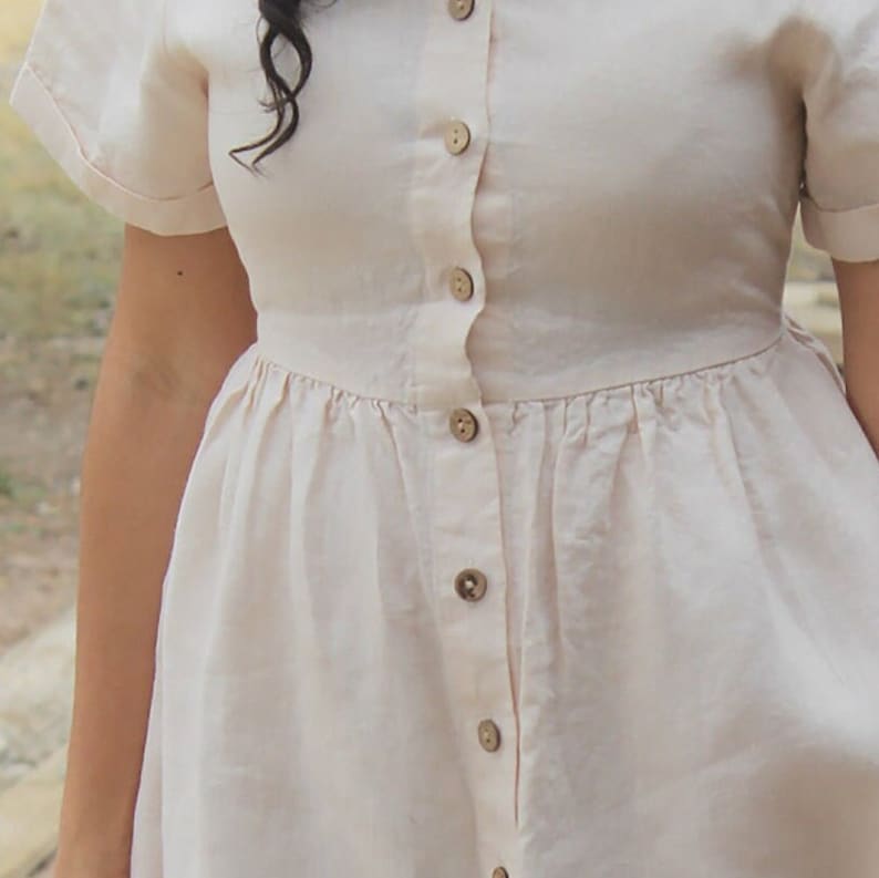May include: A white linen dress with a button-up front and a gathered skirt. The dress has short sleeves and a relaxed fit.
