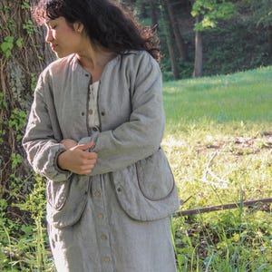 May include: A light gray, vertically striped jacket and matching button-down skirt. The jacket has large, rounded pockets and a round collar. The outfit is worn over a cream-colored top. The outfit is made of a soft, textured fabric.