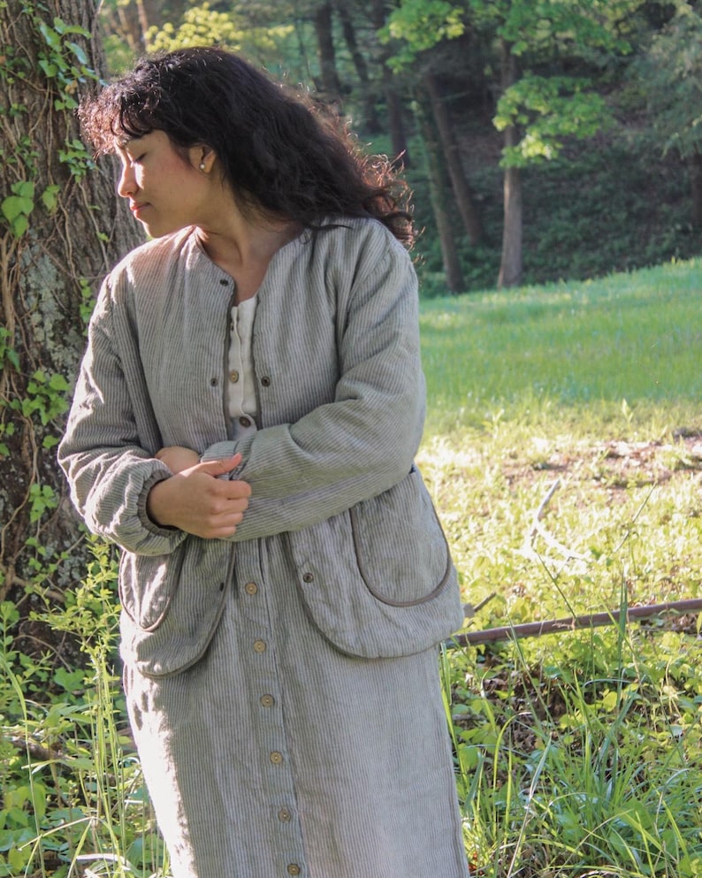 May include: A light gray, vertically striped jacket and matching button-down skirt. The jacket has large, rounded pockets and a round collar. The outfit is worn over a cream-colored top. The outfit is made of a soft, textured fabric.