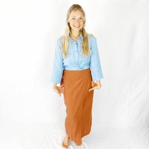 May include: A woman wearing a light blue button-down shirt and a rust-colored wrap skirt. She is smiling and looking at the camera.