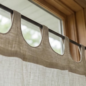 May include: A light beige curtain with a textured weave hanging from a dark metal rod. The curtain features a series of wide, integrated fabric loops at the top that slide directly onto the rod, creating a clean and simple window treatment.
