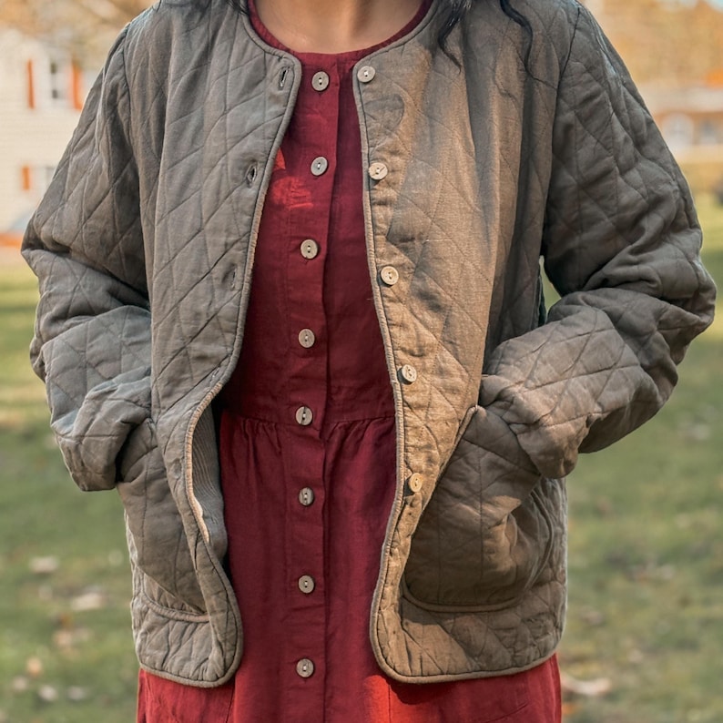 Puede incluir: Chaqueta acolchada verde oliva con cuello redondo y cierre de botones. La chaqueta tiene dos bolsillos delanteros y se lleva sobre un vestido rojo. El dise&ntilde;o acolchado de la chaqueta a&ntilde;ade textura, y el color neutro complementa el vestido.