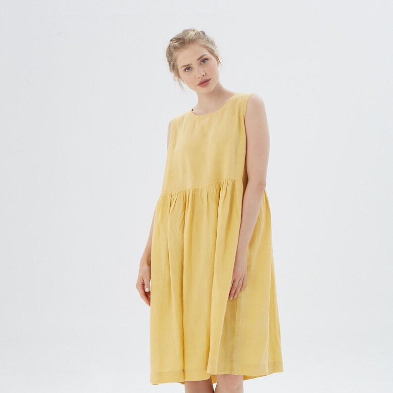 May include: A sleeveless, yellow linen dress with a gathered waist and a knee-length hem. The dress has a simple, round neckline and a relaxed fit. The fabric appears to be lightweight and breathable, suitable for warm weather.