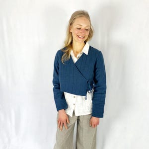 May include: A blue quilted wrap jacket with a long sleeve and a tie closure. The jacket is worn over a white button-down shirt and brown pants.