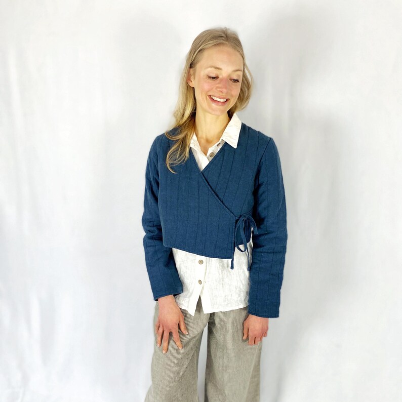 May include: A blue quilted wrap jacket with a long sleeve and a tie closure. The jacket is worn over a white button-down shirt and brown pants.