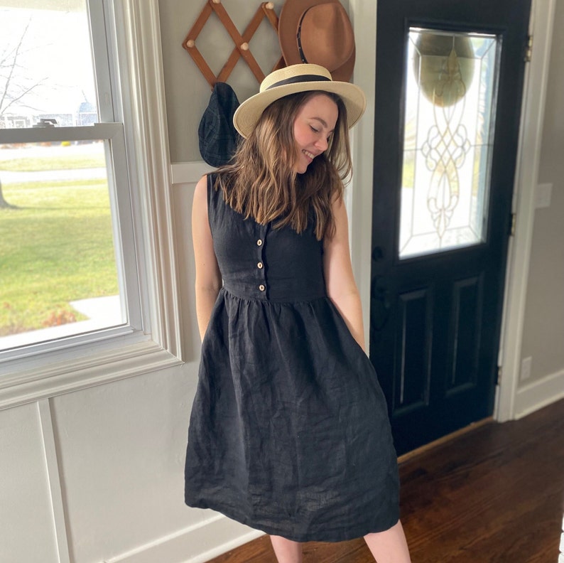 Pode incluir: Uma mulher vestindo um vestido de linho preto com bot&otilde;es na frente e um chap&eacute;u de palha. O vestido tem um corpete justo e uma saia cheia.