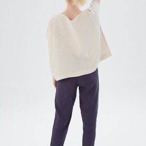 May include: A person wearing a loose-fitting, off-white linen top with a scoop neck and a pair of dark purple linen pants.